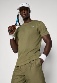 Ελαιόχρωμο βαμβακερό t-shirt με μικρό πράσινο λογότυπο, σε συνδυασμό με ταιριαστά σορτς, λευκά περιβραχιόνια και λευκό καπέλο, ενώ κρατάει ένα ρακέτα του τένις.