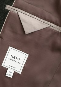 Brunt stof med en glat tekstur, der har et lys beige foer og et hvidt mærke med "NEXT TAILORING," størrelse "40R," og "SLIM FIT."