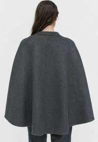 Cape en laine grise avec un col rond, design légèrement évasé et texture lisse, portée sur un pantalon sombre. La vue de dos met en avant des lignes épurées.