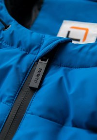 Sininen toppatakki, jossa on musta vetoketju ja vetonappi, jossa lukee "OMBRE." Takissa on hillitty tikkikuosi ja oransseilla ja mustilla yksityiskohdilla varustettu logolappu.