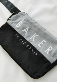 Pochette zippée noire et grise avec l'inscription « BAKER BY TED BAKER » imprimée sur le devant, posée sur un fond de tissu blanc.