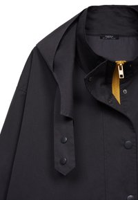 Veste de pluie noire en tissu léger, équipée d'une fermeture éclair jaune, de boutons-pression et d'une capuche avec un col structuré.