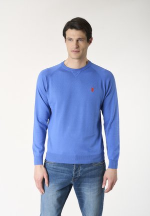 Uomo che indossa un maglione blu a maniche lunghe con scollo tondo e un piccolo emblema rosso, abbinato a jeans blu, in piedi davanti a uno sfondo semplice.