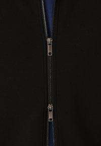 Black fabric with a double zipper revealing a glimpse of blue fabric beneath, showing metallic zipper pulls in the center.