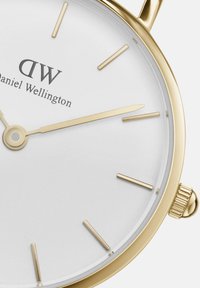 Zelta tonis rokas pulkstenis ar baltu ciparnīcu, plānām stundu atzīmēm un teksturētu kroni. Zīmola nosaukums "Daniel Wellington" izvietots virs centra.