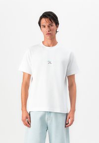 Jovem vestido com uma t-shirt branca com um pequeno gráfico azul e castanho ao centro, combinada com calções de ganga azuis claros, de pé contra um fundo branco.