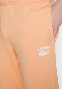 Peachfärgade bomullsbyxor i sweatpants-stil med elastisk midja, sidofickor och en vit broderad Lacoste-logotyp. Slät textur.
