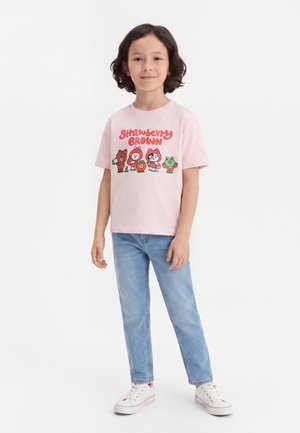 Enfant portant un t-shirt graphique Strawberry Brown rose clair, un jean bleu clair et des baskets blanches, debout devant un fond blanc uni.