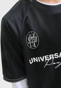 Svart idrottströja med en slät textur. Har en rund hals, grå randiga detaljer och vittryckta logotyper och text.