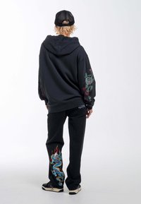 Sudadera negra con diseño de mangas florales, combinada con unos jeans negros que presentan un bordado colorido de dragón en la pierna izquierda, y zapatillas negras.