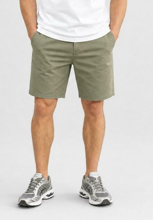 Man die olijfgroene korte broek, wit t-shirt en grijze hardloopschoenen draagt, staat met handen in de zakken tegen een effen achtergrond.