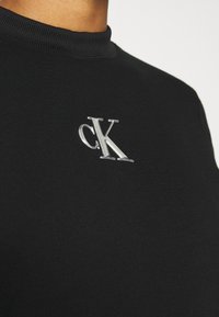 Schwarzer Sweatshirt mit Rundhalsausschnitt aus glattem Stoff, mit einem silbernen "cK"-Logo auf der Brust bestickt, und einem gerippten Kragen.