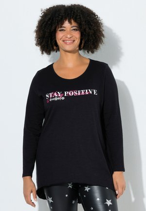 Lachende vrouw met krullend haar die een zwart shirt met lange mouwen draagt met de tekst "Stay Positive" en glanzende zwarte legging met zilveren sterren.