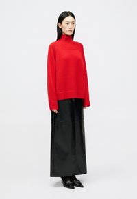 Nainen, jolla on päällään punainen korkea kaulusneule, pitkä musta nahkahame, mustat sukkahousut ja mustat kärkitossut, seisoo yksinkertaista taustaa vasten.