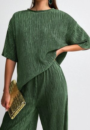 Femme portant un ensemble haut et pantalon verts texturés, tenant une pochette dorée texturée, une main sur la hanche et des boucles d’oreilles géométriques vertes visibles.