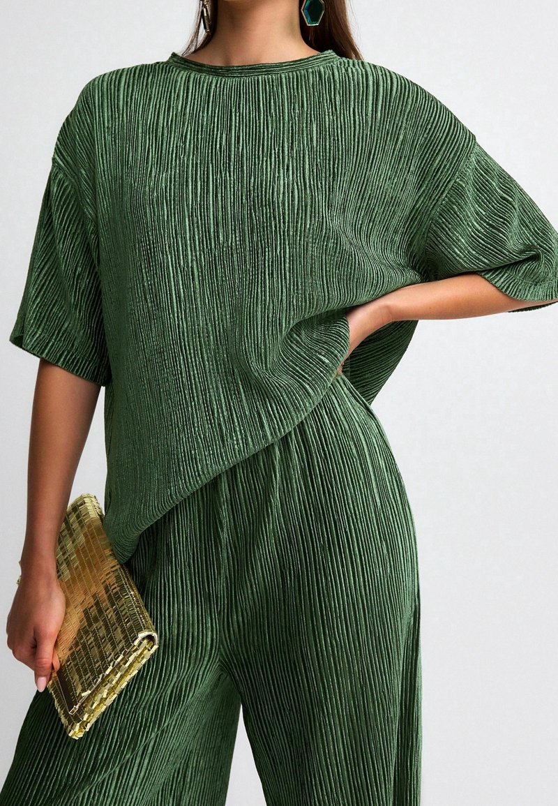 Femme portant un ensemble haut et pantalon verts texturés, tenant une pochette dorée texturée, une main sur la hanche et des boucles d’oreilles géométriques vertes visibles.