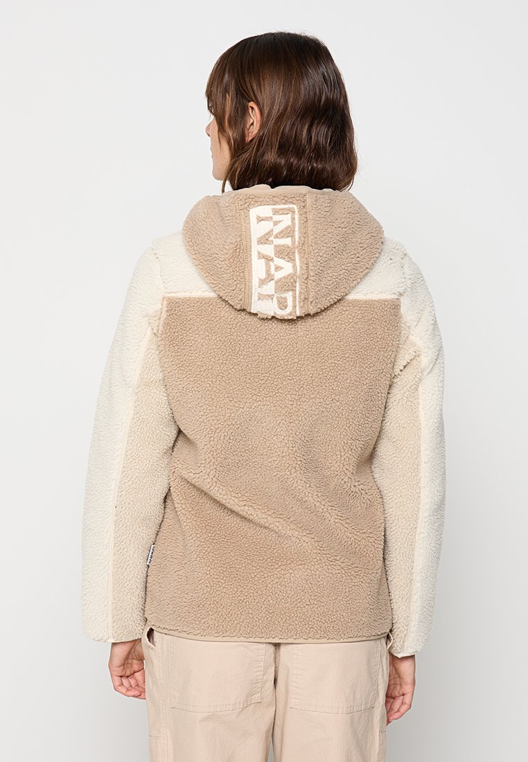 Veste en polaire beige et crème avec capuche ; présente un tissu texturé, des panneaux contrastants et un logo affiché au dos de la capuche.