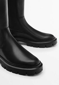 Bottes noires en cuir à cheville avec un bout arrondi, une semelle en caoutchouc épaisse et des panneaux latéraux élastiques pour un port facile. Texture lisse et design minimaliste.