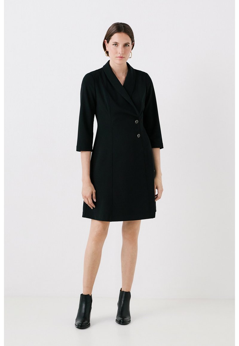 Robe blazer noire avec un col cranté, fermeture à double bouton et manches trois-quarts. Tissu lisse et silhouette ajustée. Bottines noires.