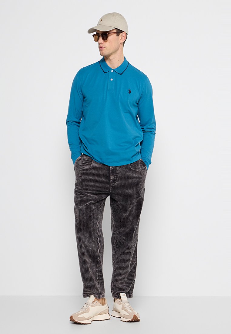 Polo a maniche lunghe azzurro con colletto, abbinato a pantaloni in velluto a coste scuri e sneakers beige. Include un cappellino leggero e occhiali da sole.