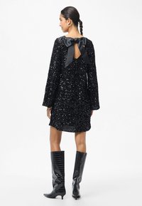 Kobieta w czarnej sukience z cekinami, z dużą satynową kokardą z tyłu i czarnych, teksturowanych kozakach do kolan, stojąca na białym tle.