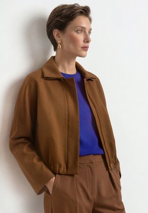 Mujer con pelo corto y castaño que lleva una chaqueta marrón, un suéter azul, pantalones marrones y pendientes dorados en forma de lágrima, de pie contra una pared blanca.