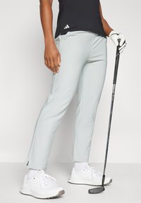 Calças de golfe azul claro com fendas laterais, combinadas com ténis brancos. Top preto texturizado e um taco de golfe segurado na mão com uma luva.