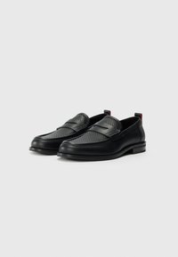 Svart lær slip-on sko med perforert overdel, flat såle, kontrastsøm og liten rød løftelapp bak. Glatt tekstur.
