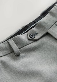 Pantalones grises con tela texturizada, con un botón negro y forro interior de lunares. Incluyen trabillas estándar para cinturón y pliegues frontales.