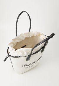 Canvas tote väska med svarta läderdetaljer, krämfärgad insida, dragsko-stängning och tryckt text på framsidan. Långa, robusta handtag.
