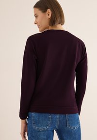 Bordeaux Sweatshirt mit rundem Halsausschnitt und langen Ärmeln, ausgestattet mit einem gerippten Saum und Bündchen, kombiniert mit blauen Jeans. Glatte Textur.