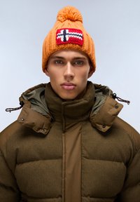 Oranžová pletená čepice s bambulí a červenou nášivkou norské vlajky. Tlustá olivově zelená prošívaná bunda s vysokým límcem a tlakacími knoflíky.
