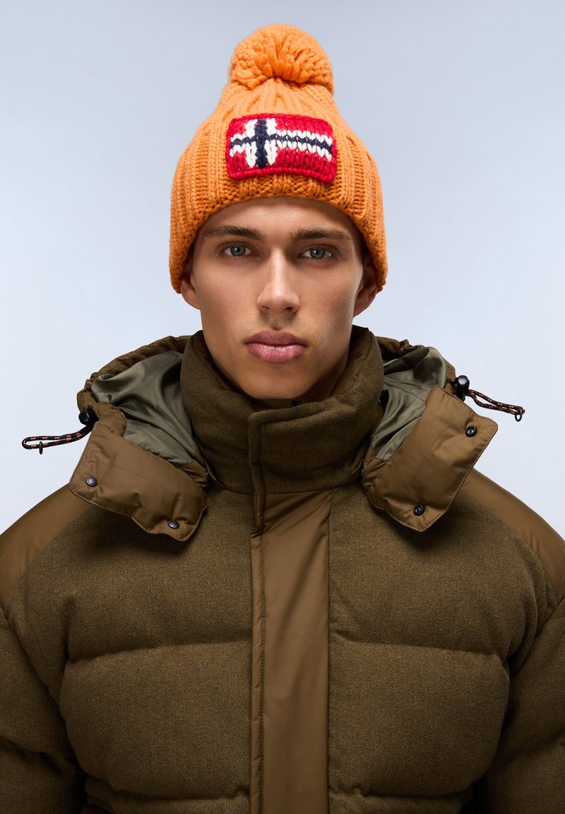 Oranžová pletená čepice s bambulí a červenou nášivkou norské vlajky. Tlustá olivově zelená prošívaná bunda s vysokým límcem a tlakacími knoflíky.