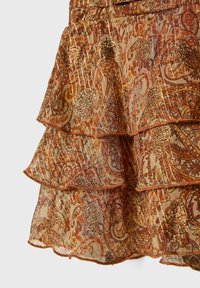Ausgestellter Stufenrock aus transparentem Stoff mit orangefarbenen und goldenen Paisleymustern, verziert mit metallischen goldenen Akzenten und einer strukturierten Oberfläche.