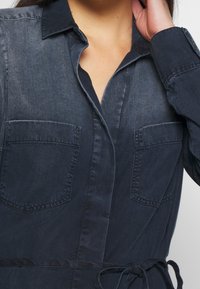 Abito camicia in denim scuro con colletto a bottoni, due tasche sul petto e vita regolabile con cintura. Tessuto liscio con dettagli di leggero sbiadimento.