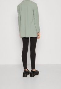 Jasnozielona marynarka z długim rękawem o luźnym kroju, zestawiona z czarnymi spodniami slim-fit i masywnymi czarnymi butami, podkreślająca gładkie faktury.