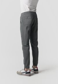 Hombre vestido con pantalones grises con cintura elástica y zapatillas blancas con detalles negros, beige y rojos, de pie sobre un fondo liso y claro.