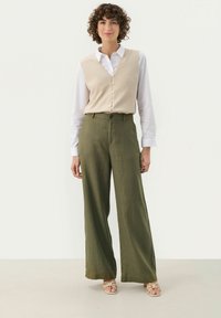 Cardigan beige con bottoni sopra una camicia bianca, abbinato a pantaloni larghi verde oliva e sandali chiari, su uno sfondo neutro.