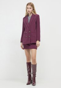 Giovane donna che indossa un completo giacca doppiopetto viola e gonna, con stivali viola lucidi alti fino al ginocchio, in piedi contro uno sfondo bianco semplice.