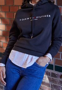 Hoodie noir avec le logo "TOMMY HILFIGER" en blanc et rouge, avec cordons, superposé sur une chemise blanche à manche à motifs, associé à un jean bleu.