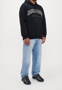 Svart huvtröja med "JACK & JONES" logotyp i vitt, tillsammans med ljusblå jeans och svarta sneakers med lila detaljer.