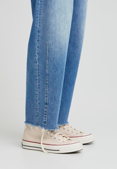 Gai zili džinsi ar platu apakšu, kopā ar bēšiem auduma sneakeriem ar baltām gumijas zolēm un sarkaniem akcentiem.