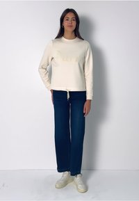 Krémová mikina s jemným logem, doplněná tmavě modrými širokými džíny a bílými teniskami. Oblečení má v pase stahovací šňůrku.