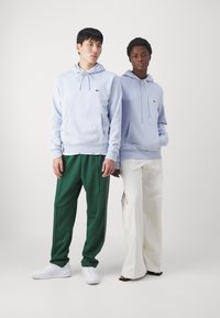 Hoodies azul claro com cordões e um bolso frontal. Um par com calças de treino verdes e ténis brancos, o outro com calças largas brancas.