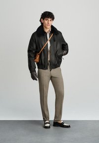 Μαύρο δερμάτινο μπομπερ μπουφάν με γούνα στο γιακά, μπεζ πουκάμισο, καστανό παντελόνι και διχρωματικά λόφερ. Κρατά μια μικρή καφέ τσάντα χιαστί.