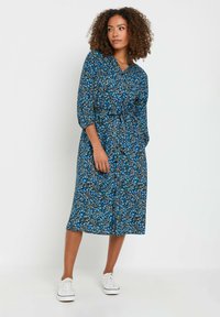 Vestido floral azul fluido con mangas tres cuartos, escote en V, cintura ajustada y dobladillo a la altura de la rodilla, combinado con zapatillas blancas.