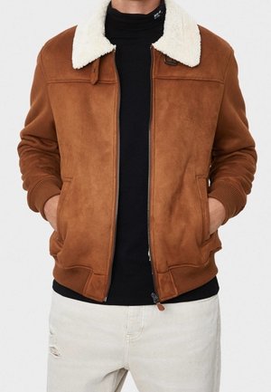 Brun ruskind bomberjakke med hvid fåreskindskrave, båret over en sort rullekrave, kombineret med lyse, revnede jeans. Hænder i lommerne.