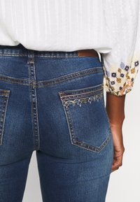 Tmavě modré denimové džíny s vyšívanými akcenty na zadních kapsách, které obsahují zlaté a barevné nitě, a mají přiléhavý střih.