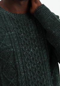 Dunkelgrüner Strickpullover mit einem Zopfmuster und gerippten Bündchen. Strukturiertes Gewebe mit verschiedenen Maschenmustern.