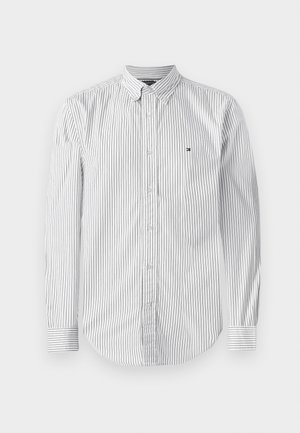 Camisa blanca de manga larga con rayas negras verticales, cuello de botón y un pequeño logo en el lado izquierdo del pecho. Material de algodón, textura suave.